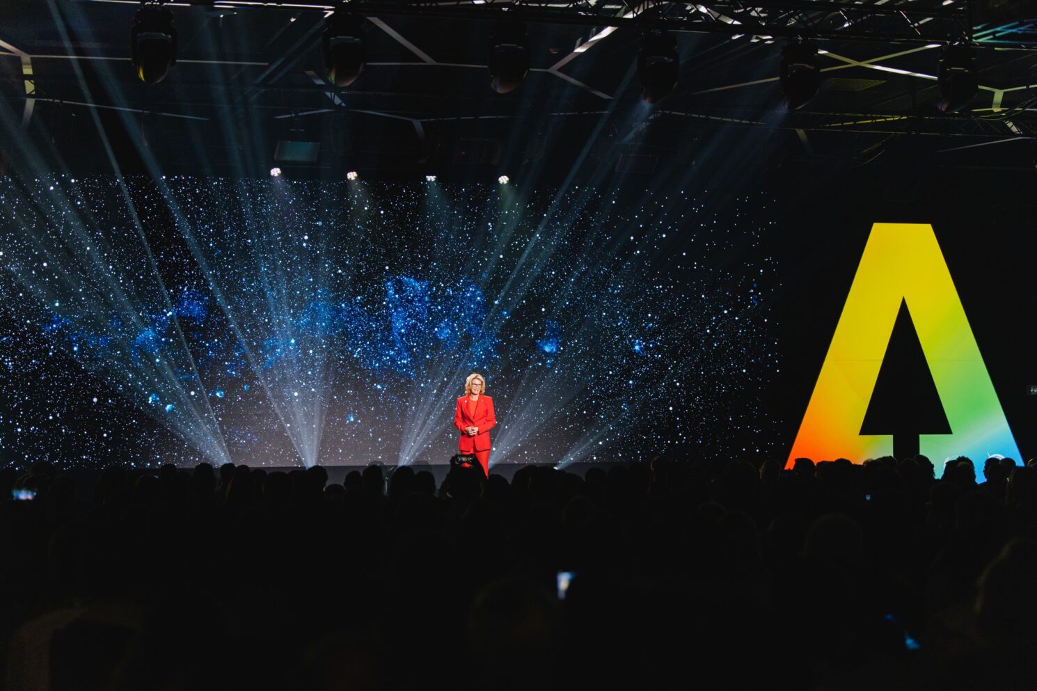 A large-scale keynote moment during the AI for Good – Global AI Conference. A speaker dressed in a striking red suit stands center stage, addressing a large audience seated in the foreground. Behind her, an expansive LED screen displays a cosmic, star-filled visual, with beams of stage light radiating outward to create a futuristic and immersive atmosphere. To the right of the stage, a monumental letter “A” in a vibrant gradient reinforces the event’s strong visual identity. The combination of advanced lighting design, digital scenography, and a focused presenter highlights the conference’s emphasis on innovation, technology, and the positive impact of artificial intelligence on society.