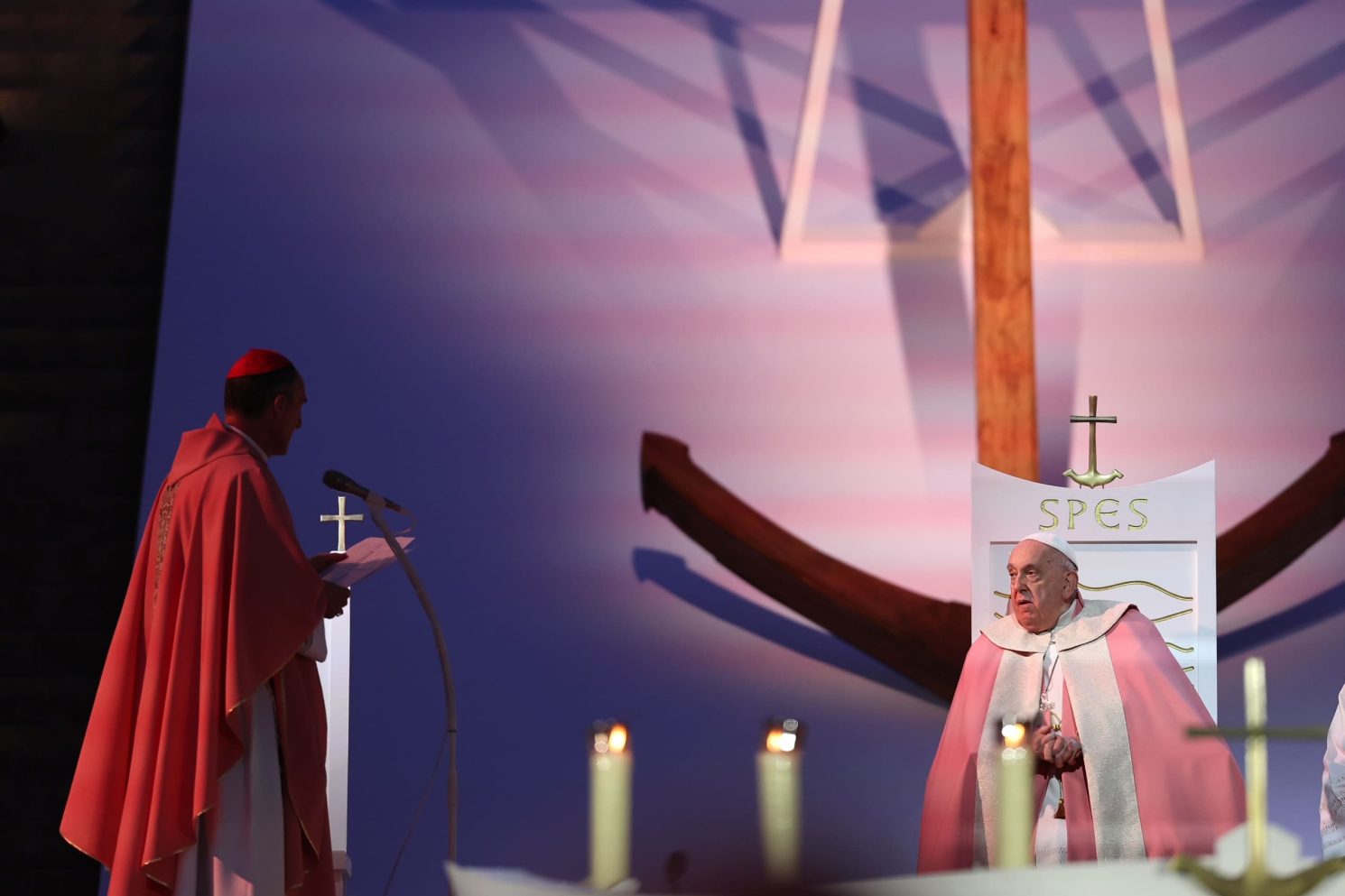 A solemn moment captured during Pope Francis’ visit to Corsica, showing the Pope seated on a ceremonial chair marked “SPES,” facing a senior clergyman dressed in red liturgical vestments who is reading from a lectern. The scene is set on a large, carefully designed stage dominated by a monumental wooden cross in the background. Soft, atmospheric lighting casts symbolic shadows across the backdrop, while candles in the foreground reinforce the sacred and contemplative tone. The composition conveys authority, reverence, and the spiritual significance of this major public religious event.