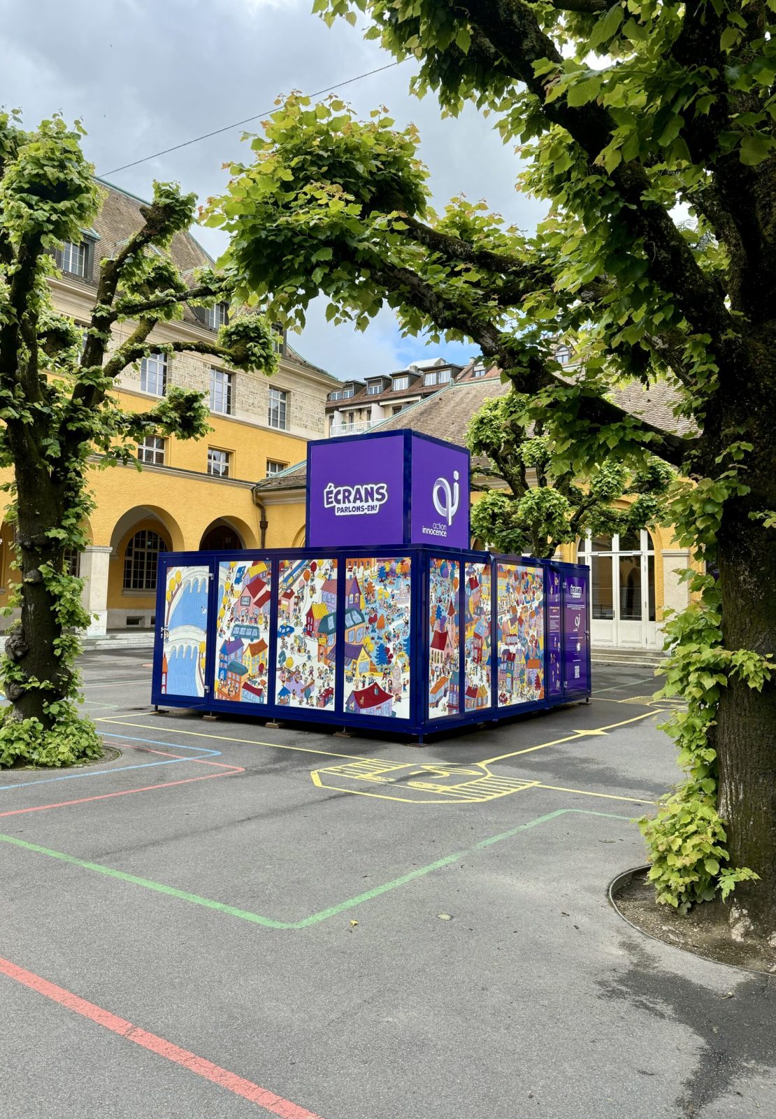 The Action Innocence traveling educational pavilion installed in an outdoor school courtyard, featuring colorful illustrated panels and branded signage, integrated into the environment beneath surrounding trees.