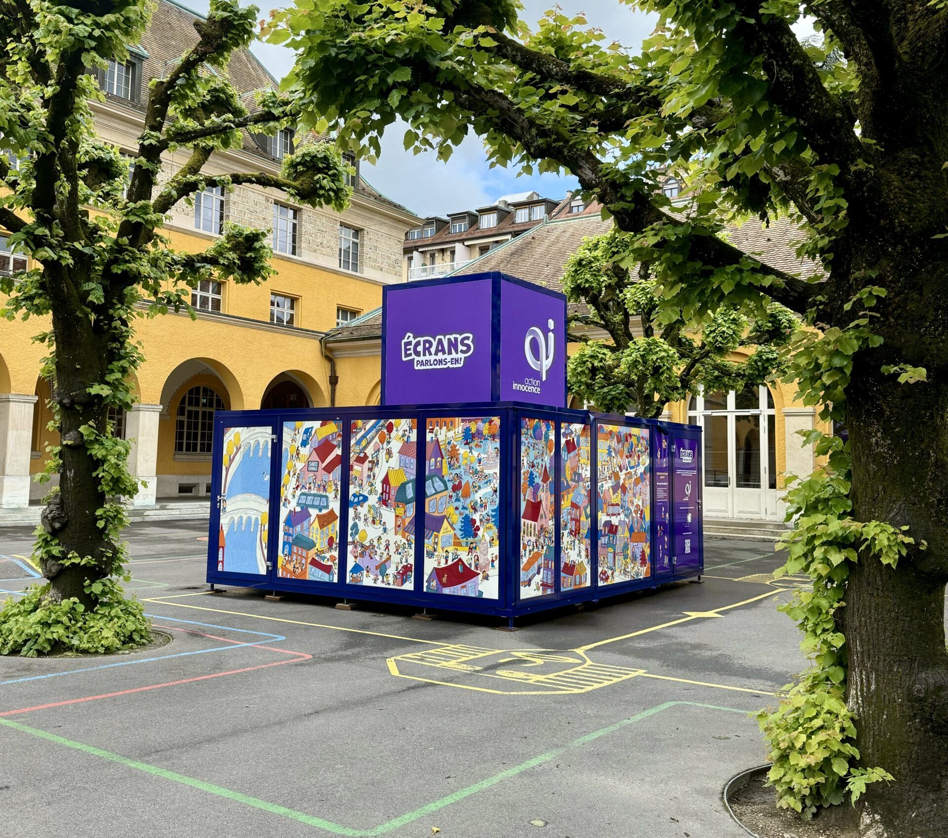 The Action Innocence traveling educational pavilion installed in an outdoor school courtyard, featuring colorful illustrated panels and branded signage, integrated into the environment beneath surrounding trees.