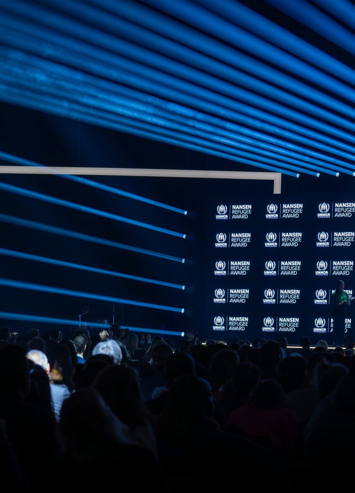 Wide view of the stage and audience during the Nansen Refugee Award – Hybrid Ceremony, with blue light beams, a branded UNHCR backdrop, and speakers addressing the crowd.