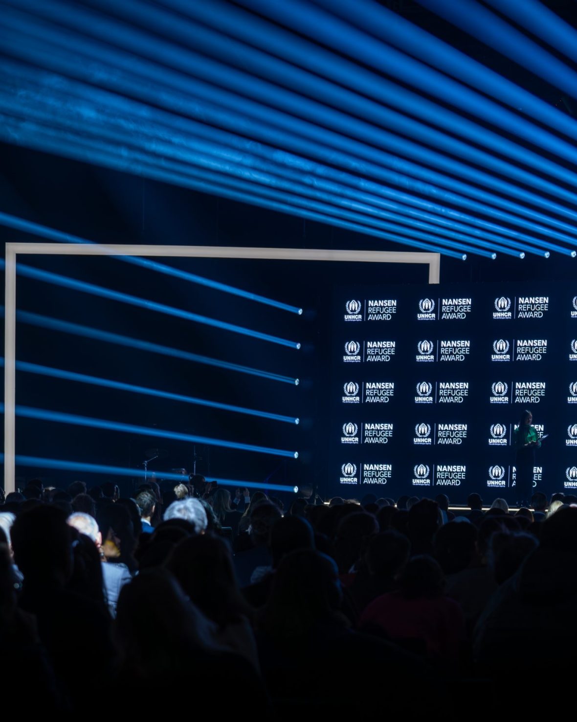 Wide view of the stage and audience during the Nansen Refugee Award – Hybrid Ceremony, with blue light beams, a branded UNHCR backdrop, and speakers addressing the crowd.