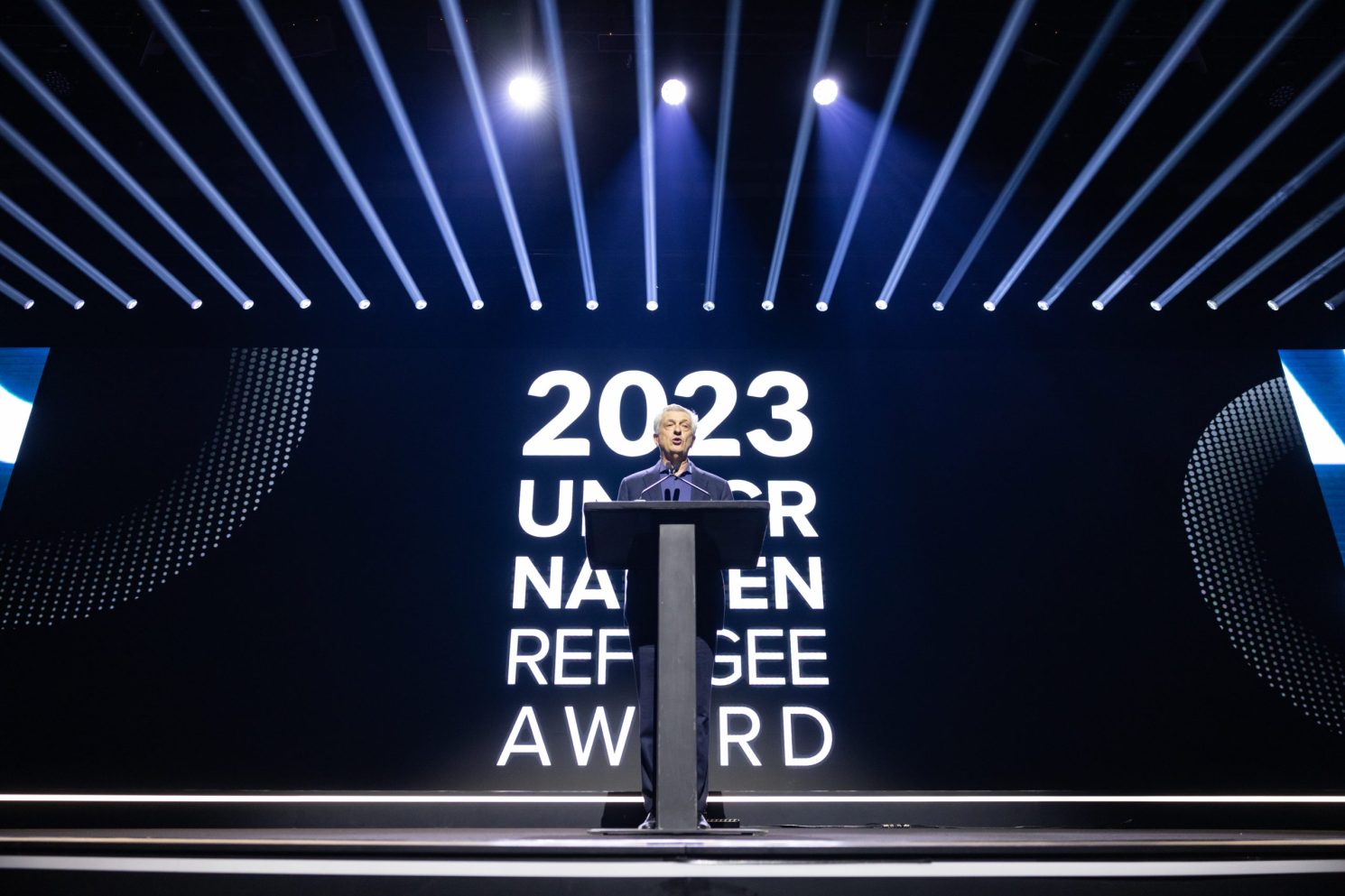 Speaker standing at a podium on a large stage during the Nansen Refugee Award – Hybrid Ceremony, with dramatic lighting and a screen displaying “2023 UNHCR Nansen Refugee Award” behind them.