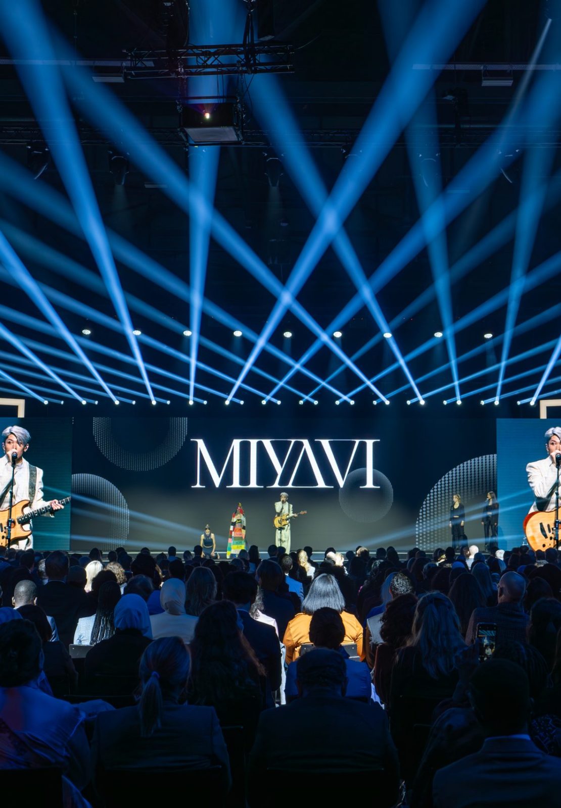 Live concert performance on a large stage with blue light beams during the Nansen Refugee Award – Hybrid Ceremony, featuring a guitarist singing at center stage and an audience seated in front of illuminated screens.