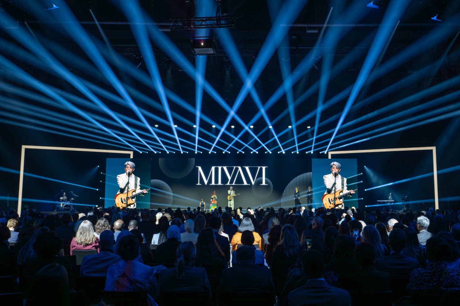 Live concert performance on a large stage with blue light beams during the Nansen Refugee Award – Hybrid Ceremony, featuring a guitarist singing at center stage and an audience seated in front of illuminated screens.