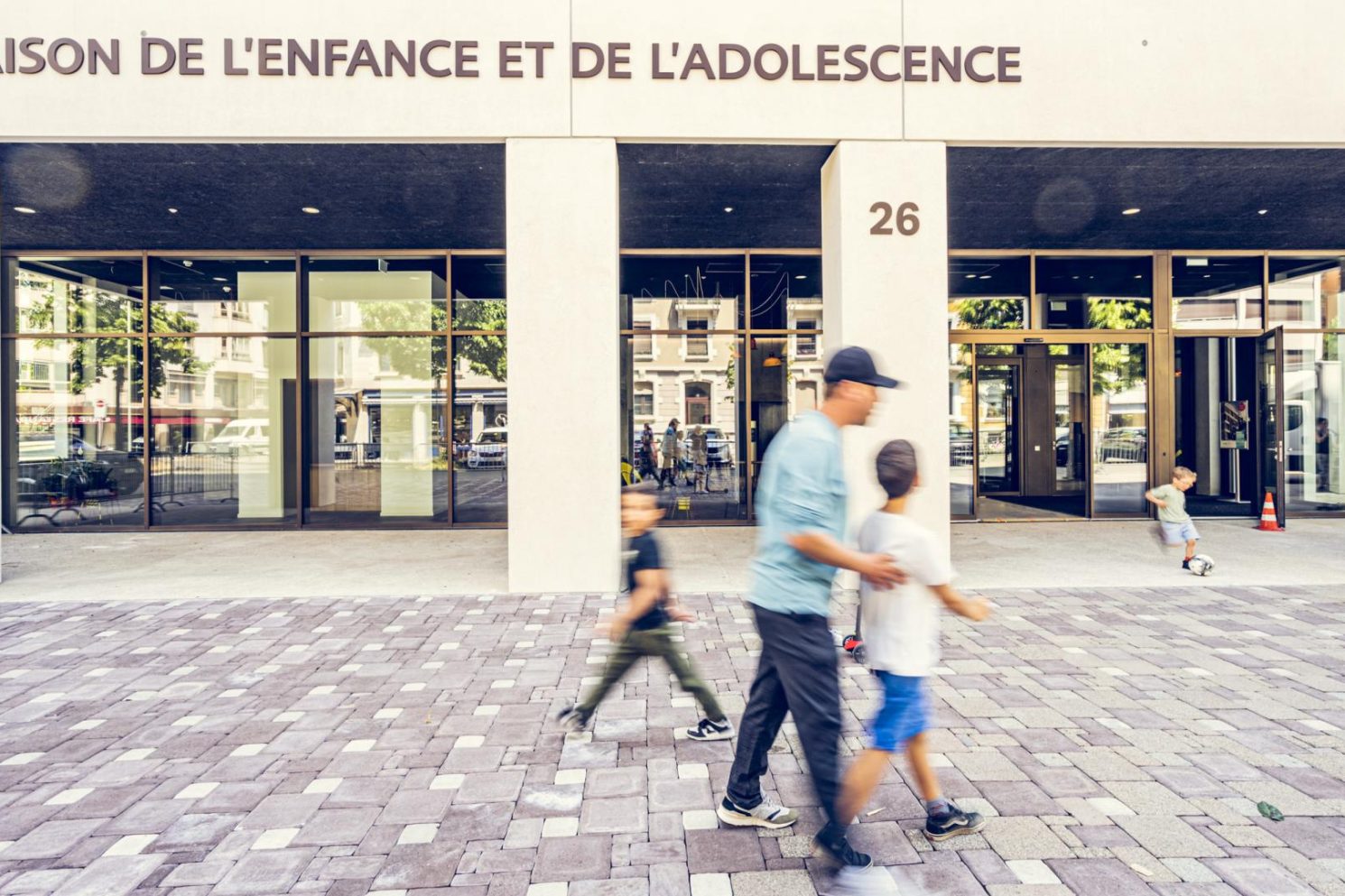 Front view of the Maison de l’Enfance et de l’Adolescence building during the MEA – Opening of MEA at Geneva, with children and an adult walking across the paved area in front of the glass entrance.