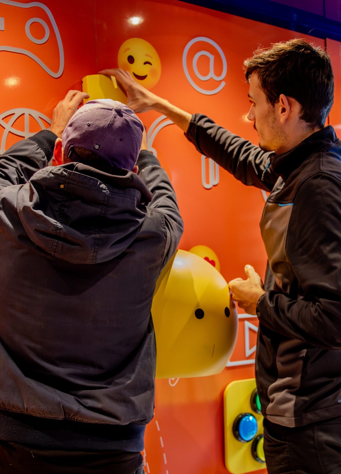 Two team members install and adjust interactive elements on a brightly colored wall, featuring digital icons and emotive symbols, as part of the hands-on setup of the Action Innocence traveling educational pavilion.