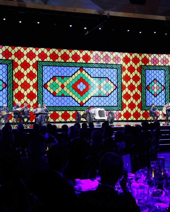 Wide stage view with live musicians, a classic race car, and a large colorful patterned backdrop during the FIA – Motorsport Award Ceremony.