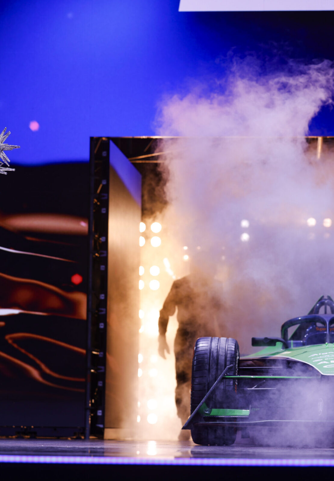 Award winner holding a trophy on stage beside a revealed race car with smoke effects during the FIA – Motorsport Award Ceremony.