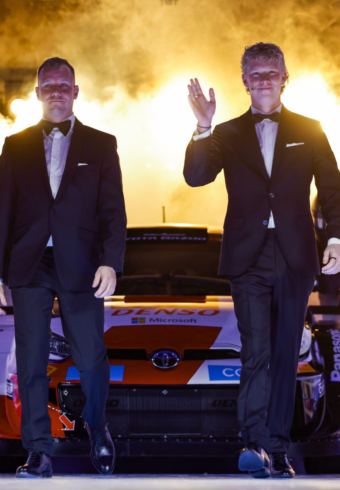 Two award winners walking on stage in front of a race car with dramatic lighting effects during the FIA – Motorsport Award Ceremony.
