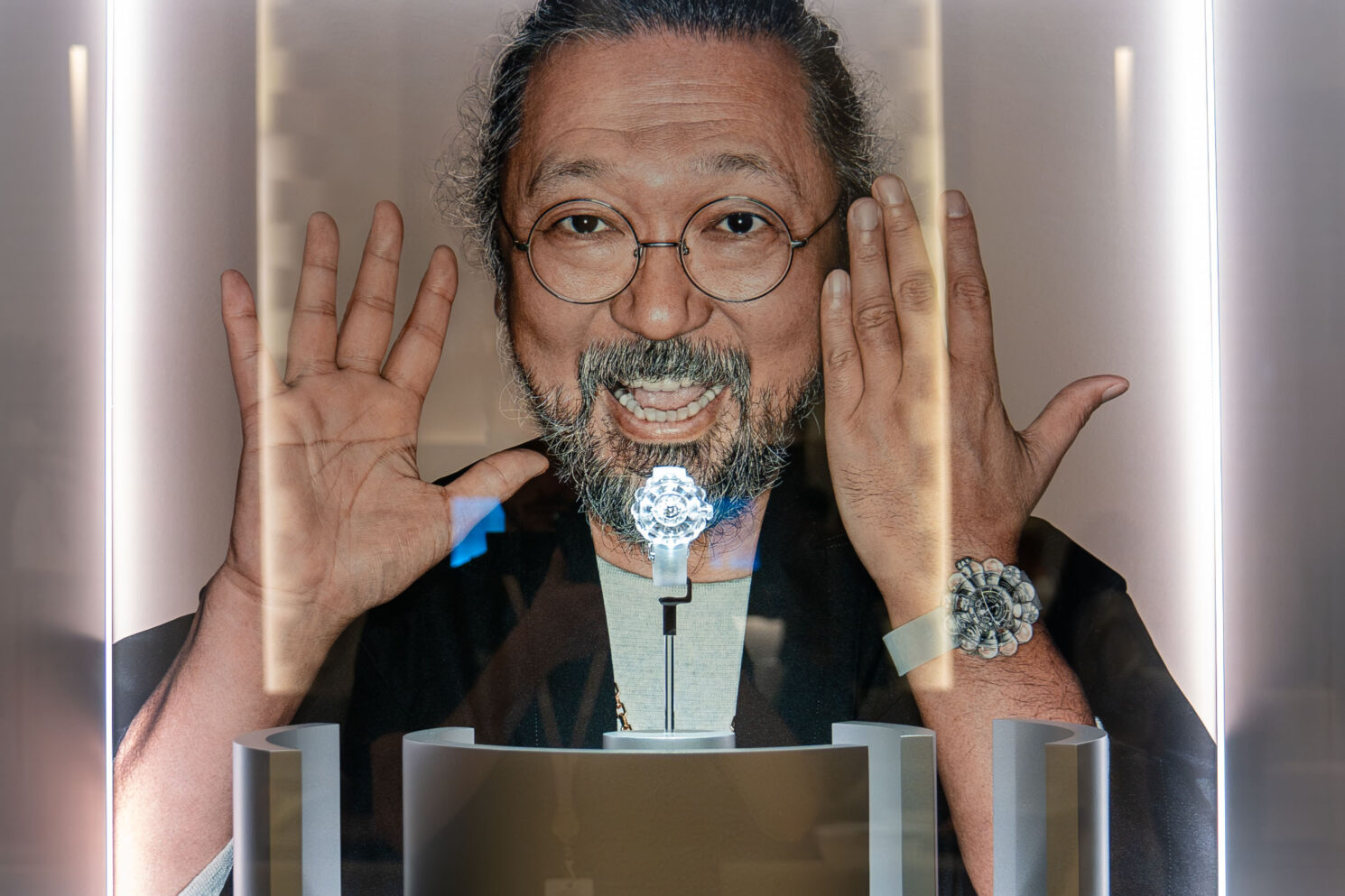 Portrait displayed behind a watch showcase, with a smiling man framed by illuminated panels and a timepiece presented in the foreground during Watches & Wonders – Geneva International Watchmaking Exhibition.