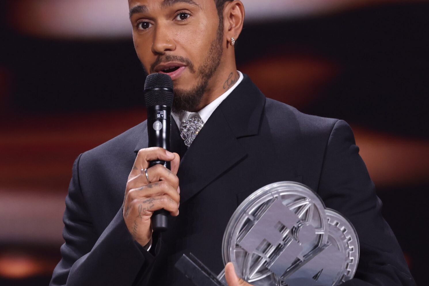 Award recipient speaking into a microphone while holding a trophy on stage during the FIA – Motorsport Award Ceremony.