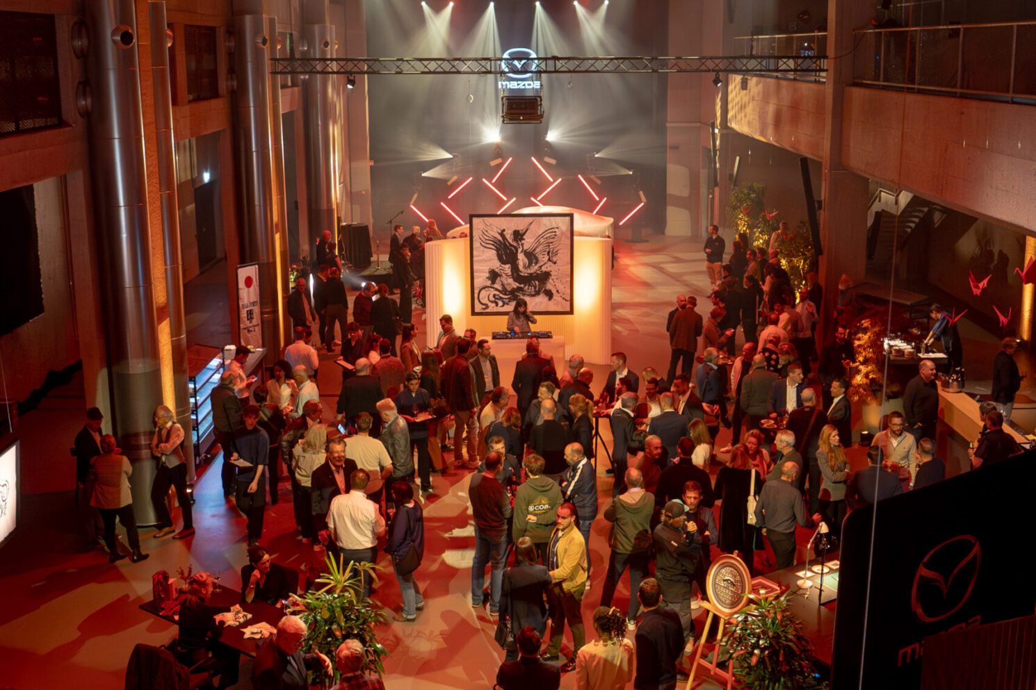 Guests gathering in a large industrial venue with dramatic red lighting during the Mazda – Cinematic reveal of the Mazda 6E.