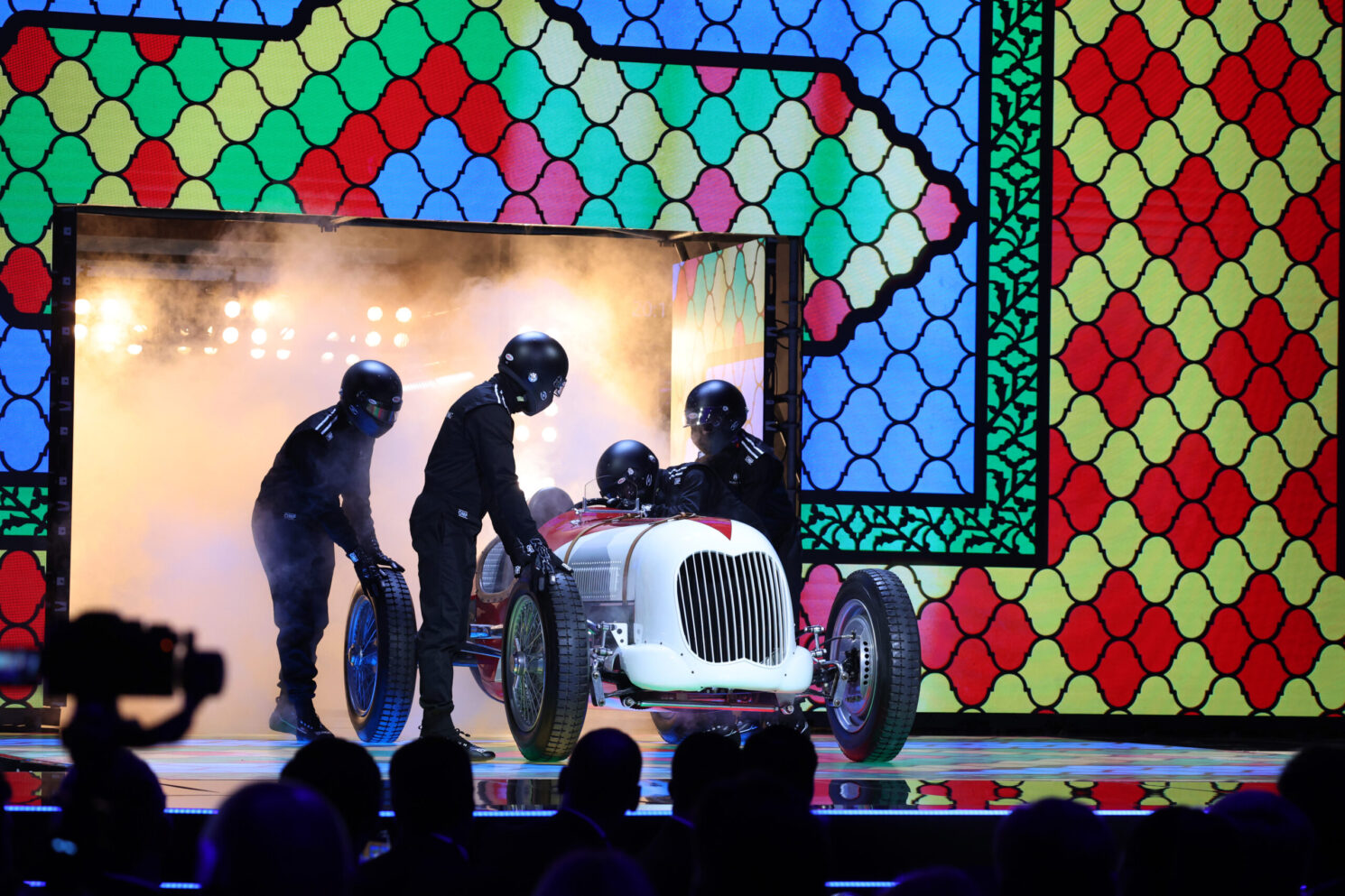 Classic race car revealed on stage by crew members during the FIA – Motorsport Award Ceremony, with theatrical lighting and a colorful patterned backdrop.