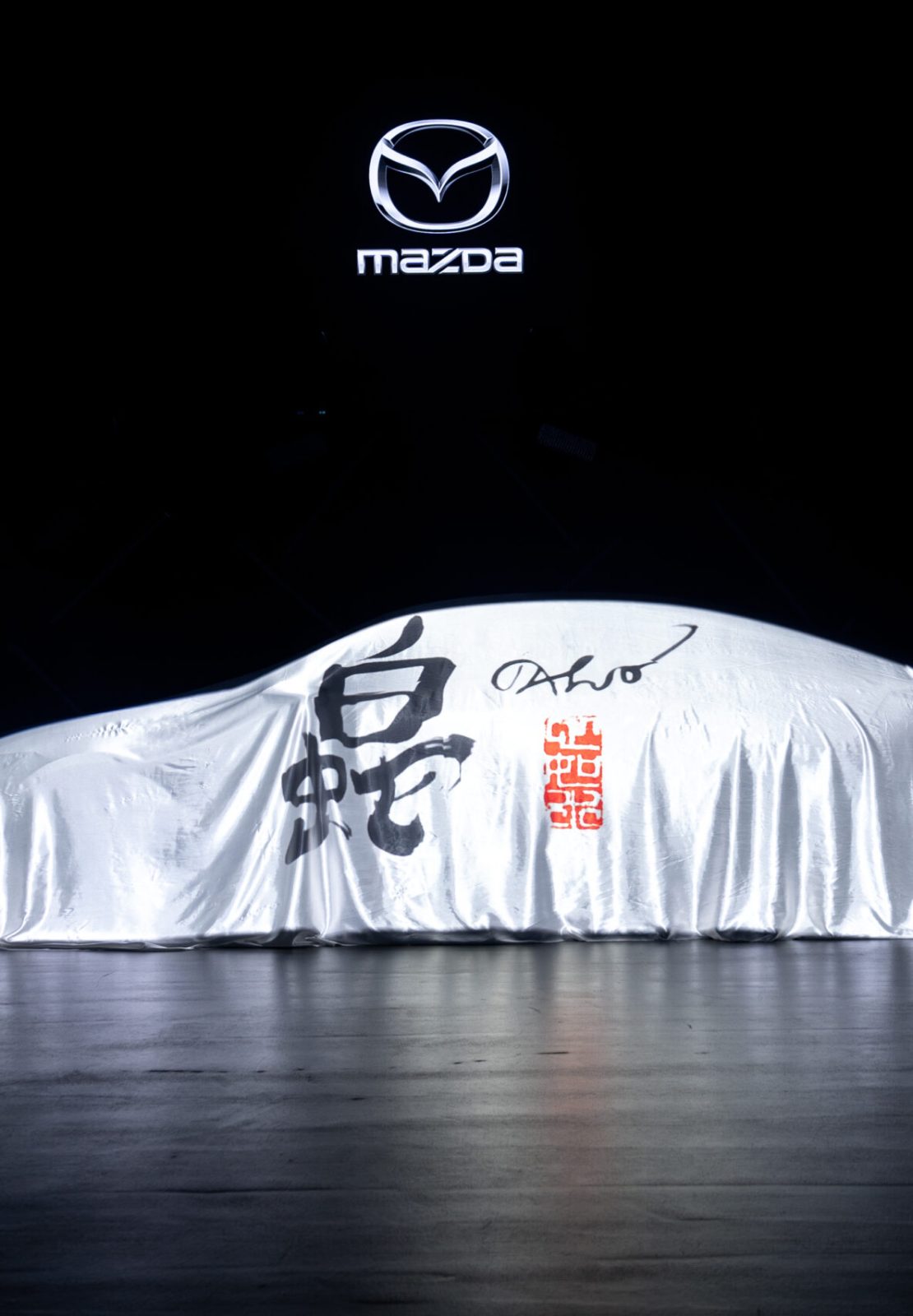 Covered car on stage beneath the Mazda logo during the Mazda – Cinematic reveal of the Mazda 6E.